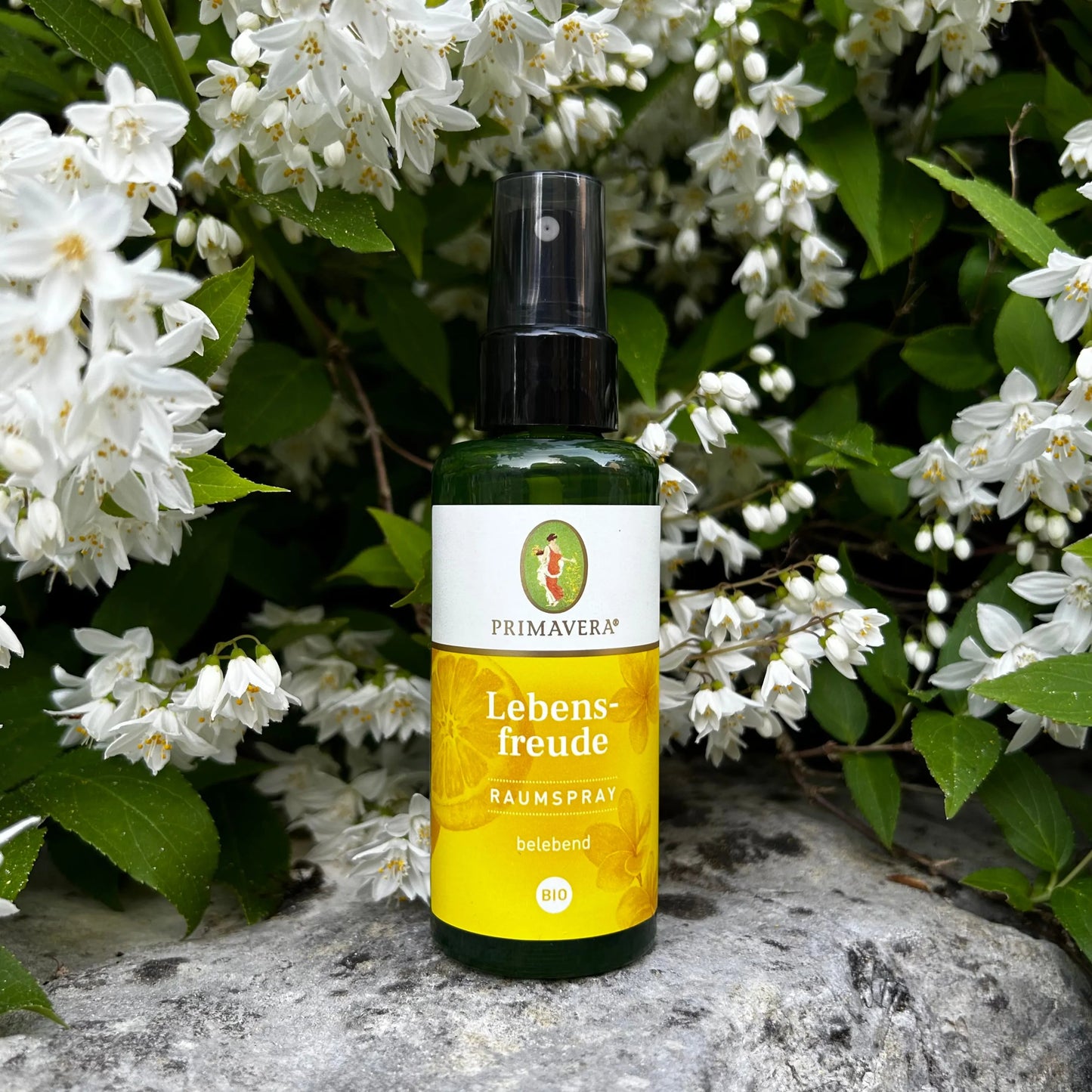 Der Lebensfreude Raumspray steht auf einem Stein vor schönen weissen Blumen.