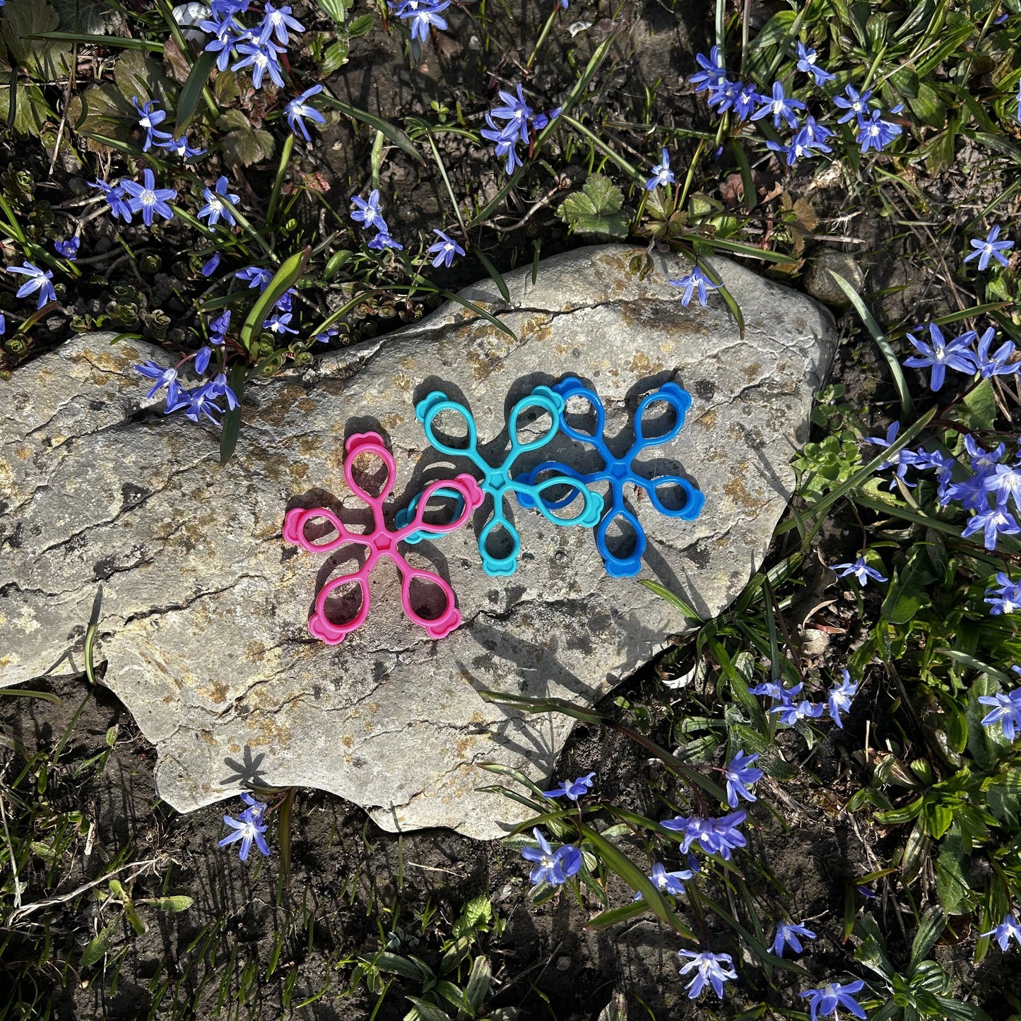 Drei Fingertrainer in türkis, rosa und blau liegen auf einem flachen grauen Stein in mitten blauer Blumen