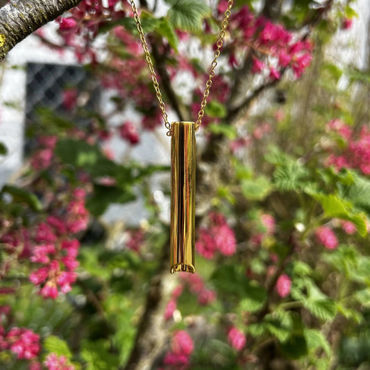 Halskette mit kurzem Röhrchen in gold hängt an einem Baum mit rosa Blüten.