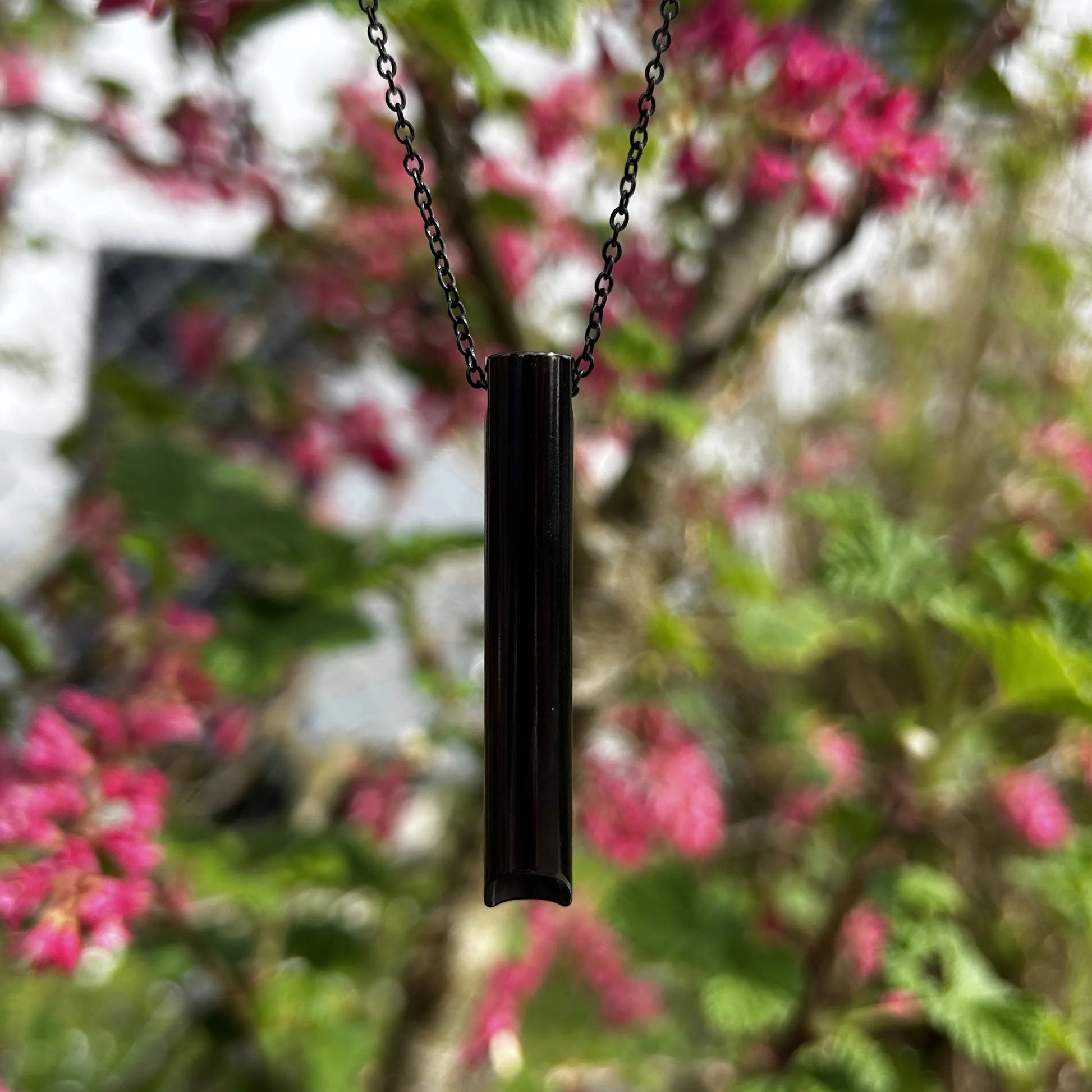 Halskette mit kurzem Röhrchen in schwarz hängt an einem Baum mit rosa Blüten.