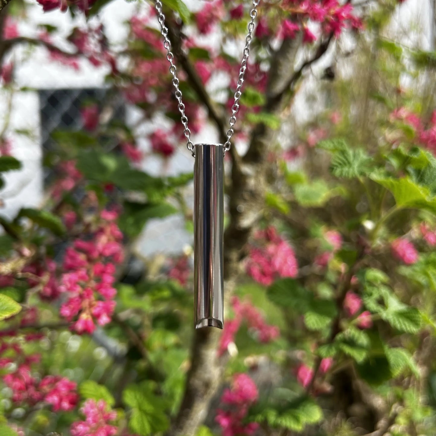Halskette mit kurzem Röhrchen in silberhängt an einem Baum mit rosa Blüten.