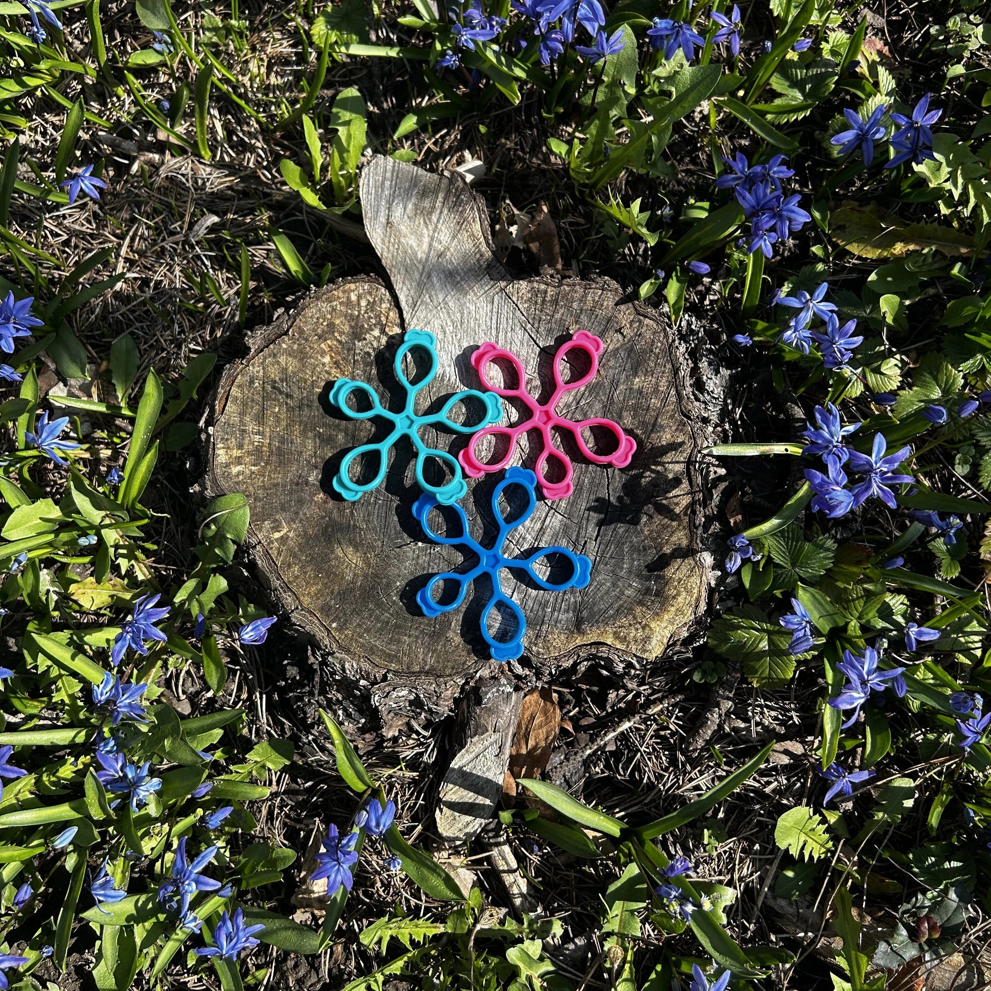 Drei Fingertrainer in türkis, rosa und blau liegen auf einem Holzstrunk in mitten blauer Blumen