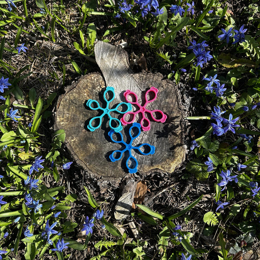 Drei Fingertrainer in türkis, rosa und blau liegen auf einem Holzstrunk in mitten blauer Blumen