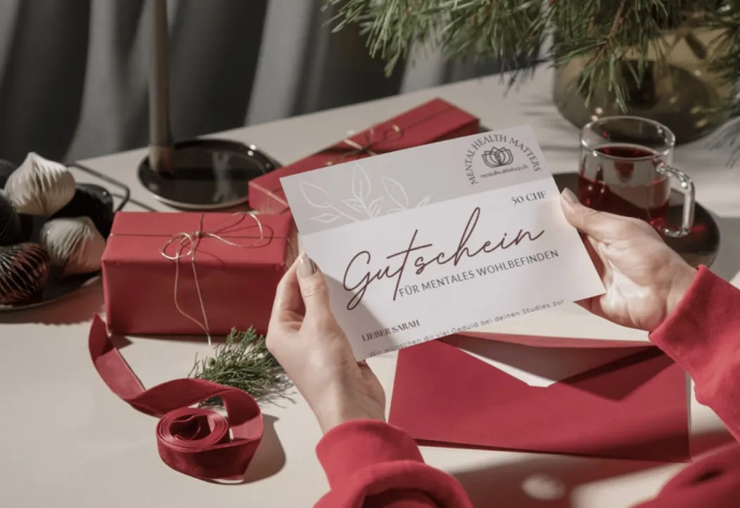 Gutschein wird in beiden Händen gehalten während die Person an einem Tisch sitzt, welcher rot eingepackte Geschenke darauf hat.